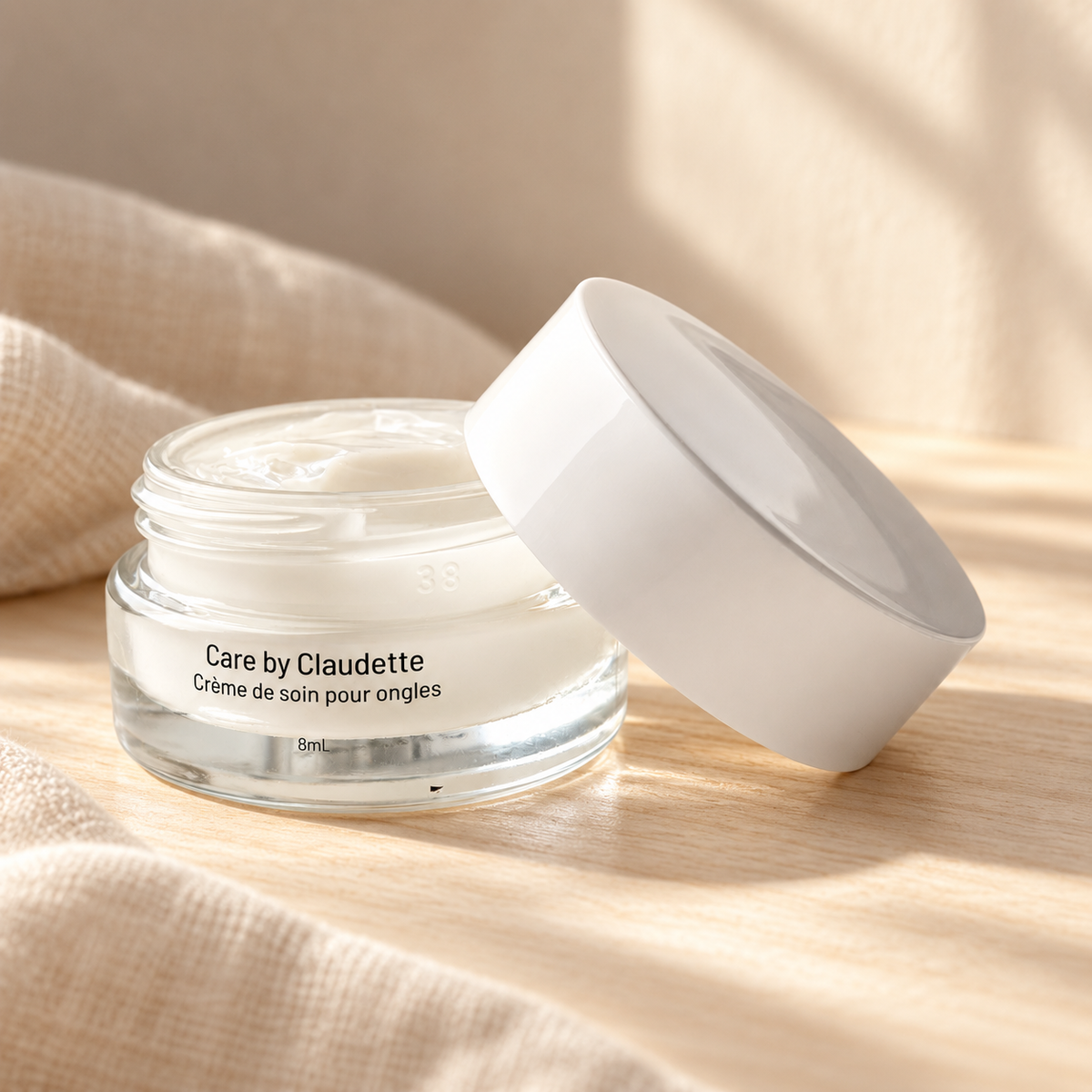 Crème de soin pour ongles Care by Claudette ouverte, mise en scène sur bois clair avec tissu beige et lumière naturelle.