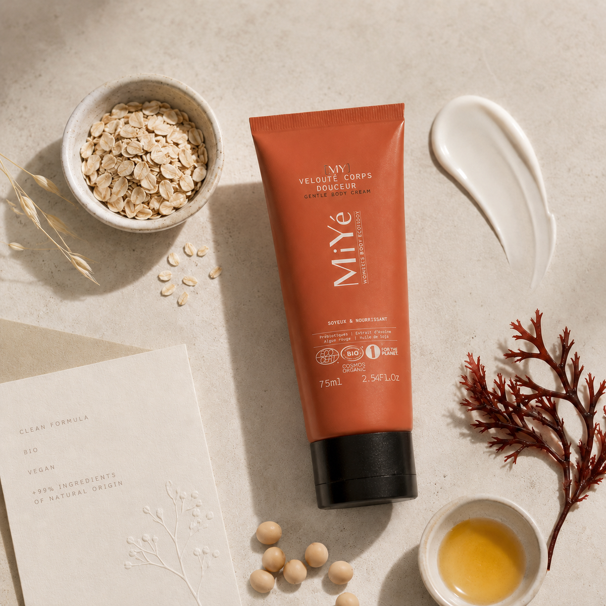 Flat lay du tube Miyé Velouté Corps Douceur entouré d’avoine, algue rouge, huile végétale, grains de soja et carte clean formula, pour illustrer une formule bio, vegan et d’origine naturelle.