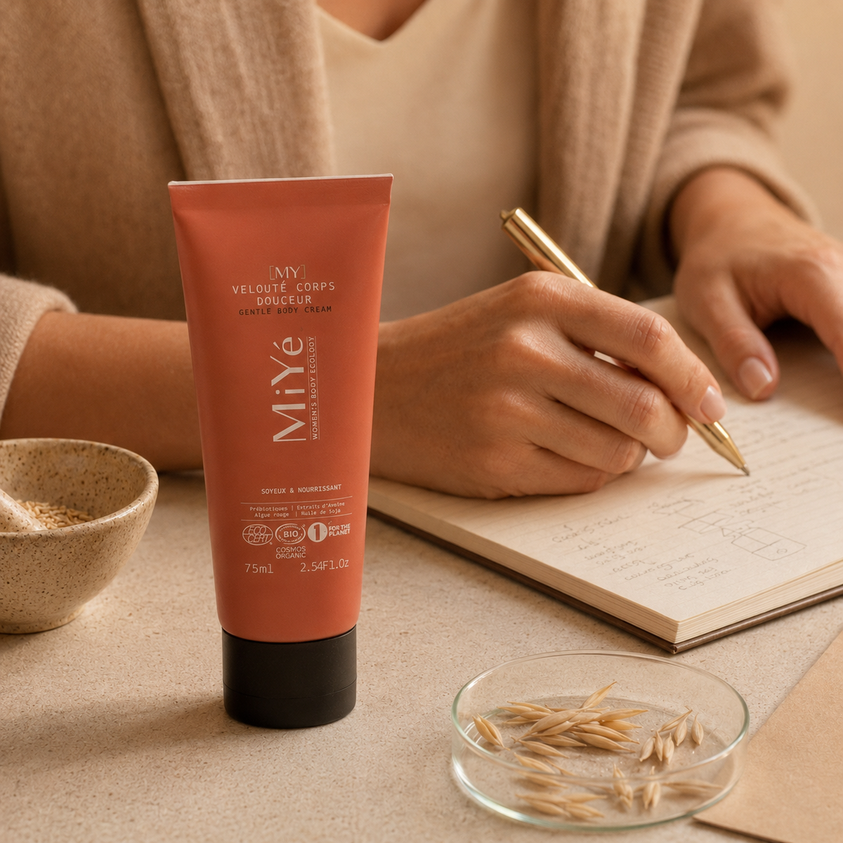 Tube Miyé Velouté Corps Douceur posé sur un bureau beige, avec carnet de formulation, main écrivant au stylo doré, bol en céramique et avoine, dans une ambiance skincare experte, douce et premium.